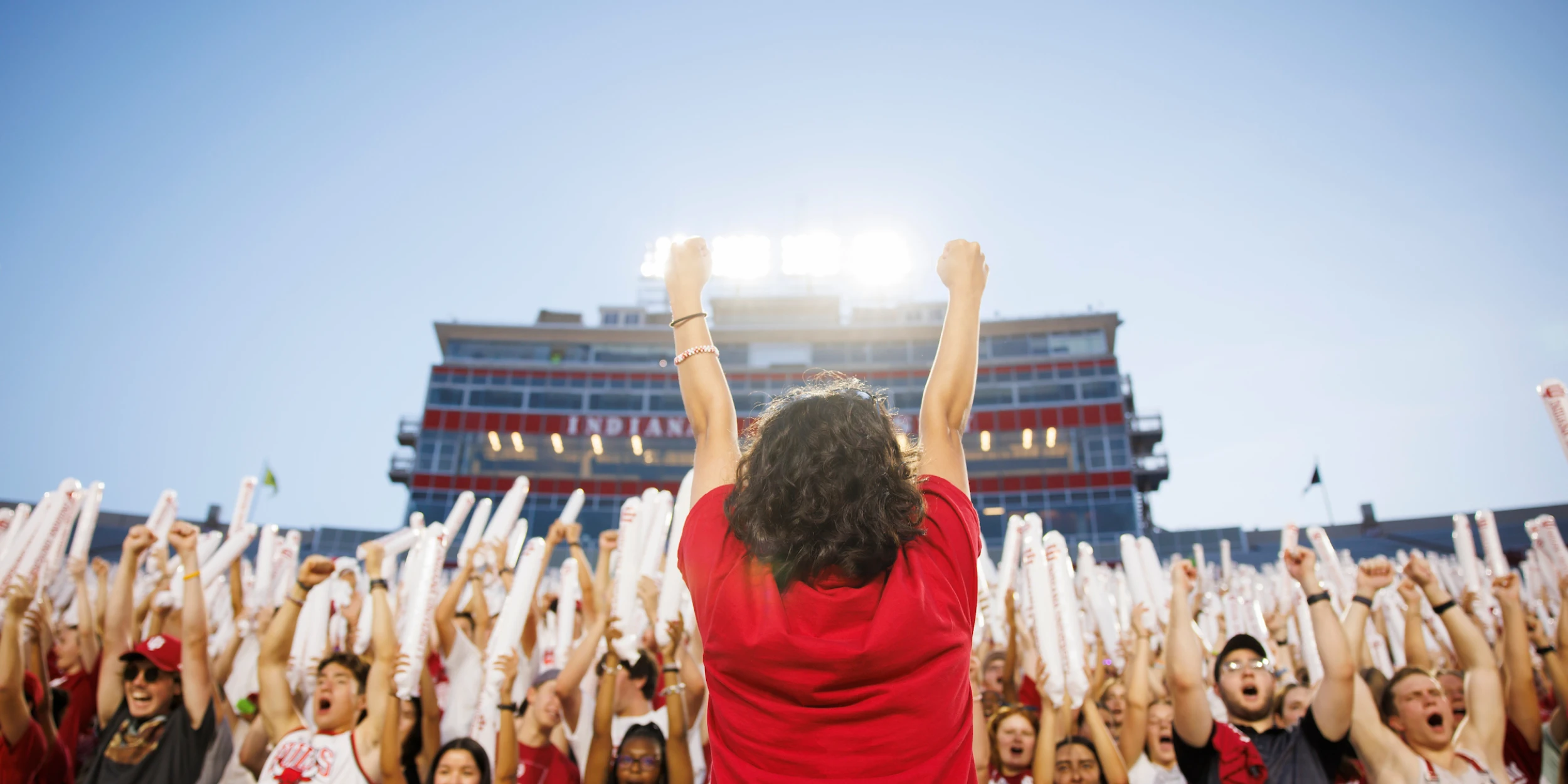 A crowd of fans, viewed from behind, cheer with their arms raised. The central figure, wearing a red shirt, has their back to the viewer and arms outstretched with fists clenched in triumph. The crowd holds white foam tubes. In the background, a large stadium building with 'INDIANA' visible on its facade stands under a bright sky.